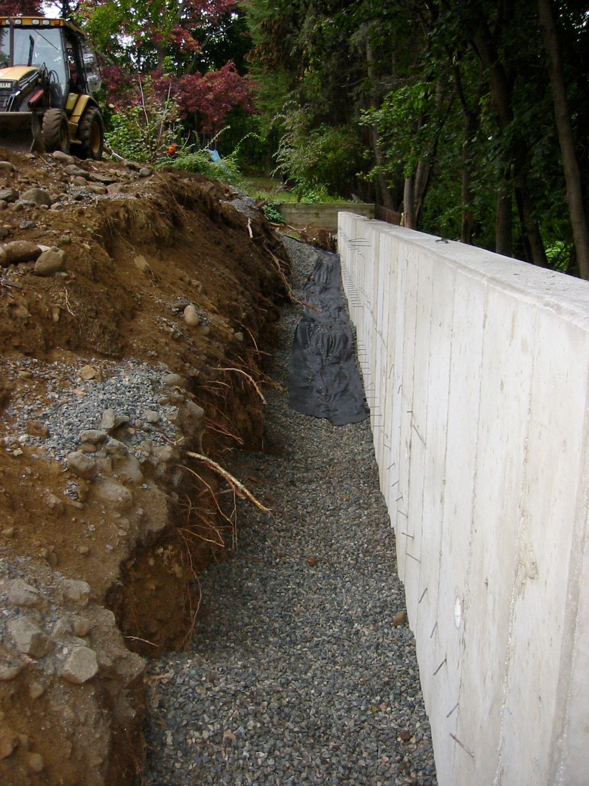Concrete retaining wall under construction next to gravel and soil. Tractor in the background.