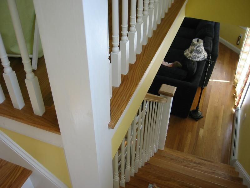 Staircase with white spindles and wooden steps, leading to a living room with a black sofa and hardwood floors.