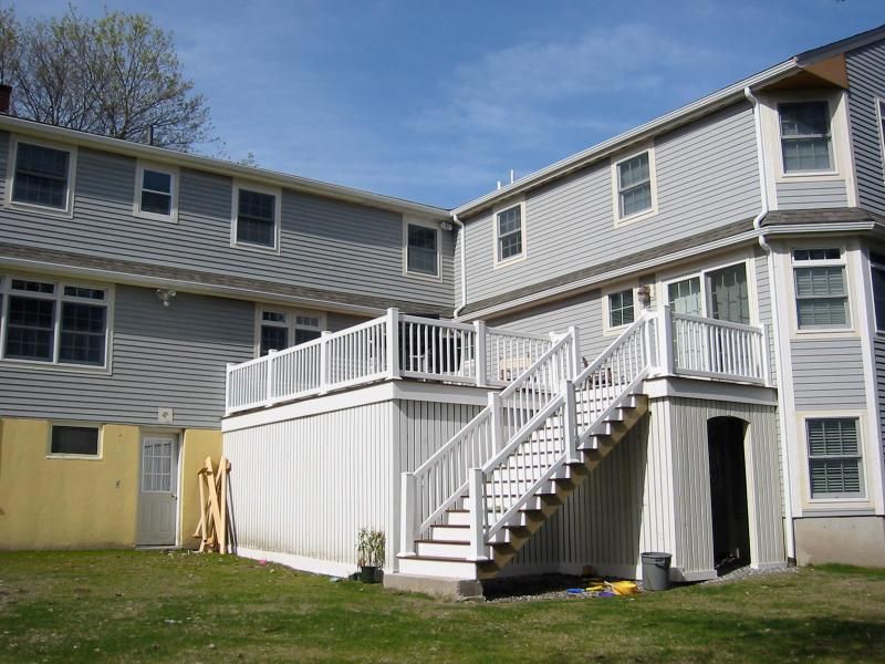 Exterior view of a two-story house with a white wooden deck and stairs. Gray siding, windows, and a grassy lawn.
