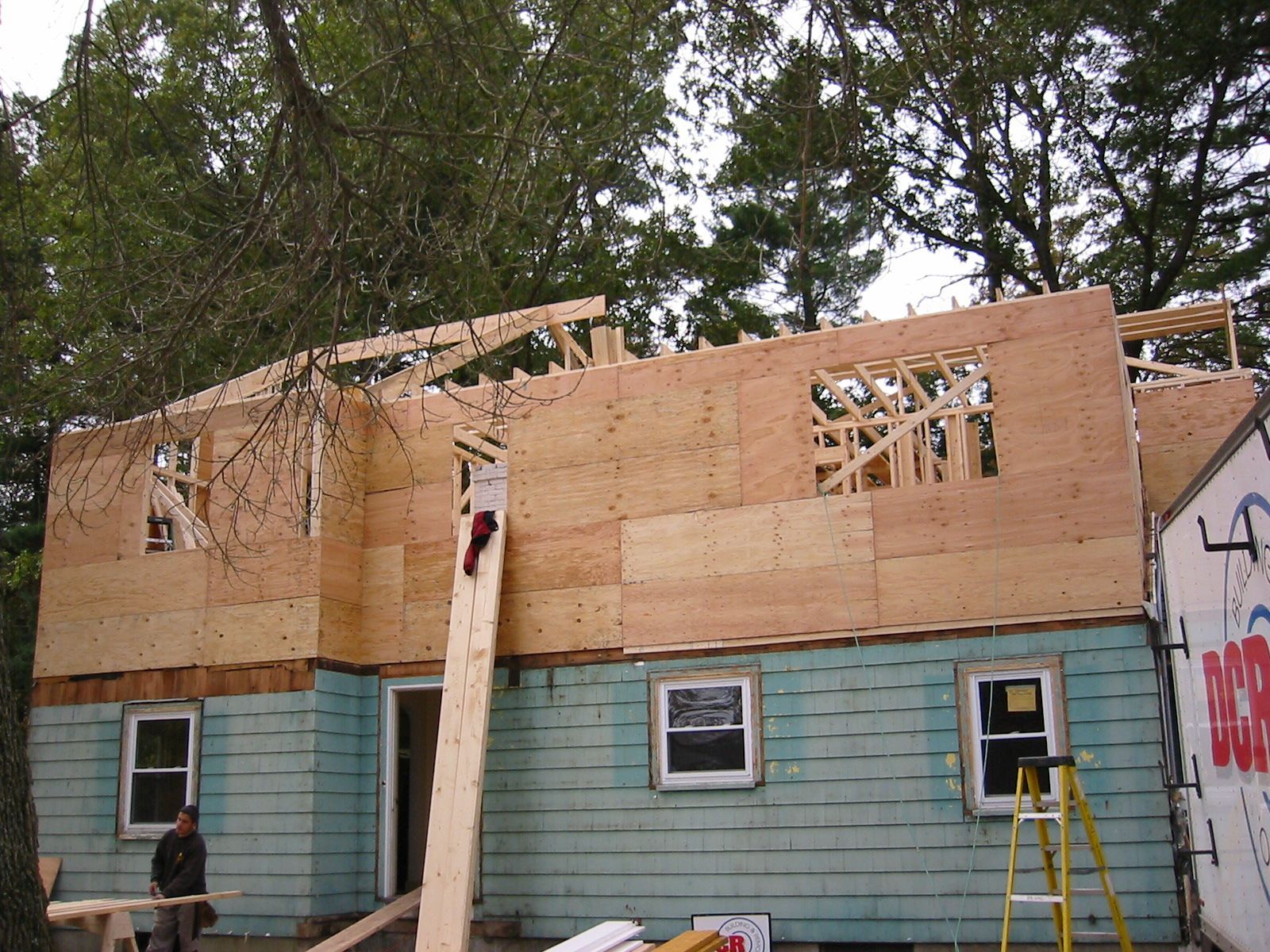 House under construction; plywood siding on upper level, teal siding below; person near base.