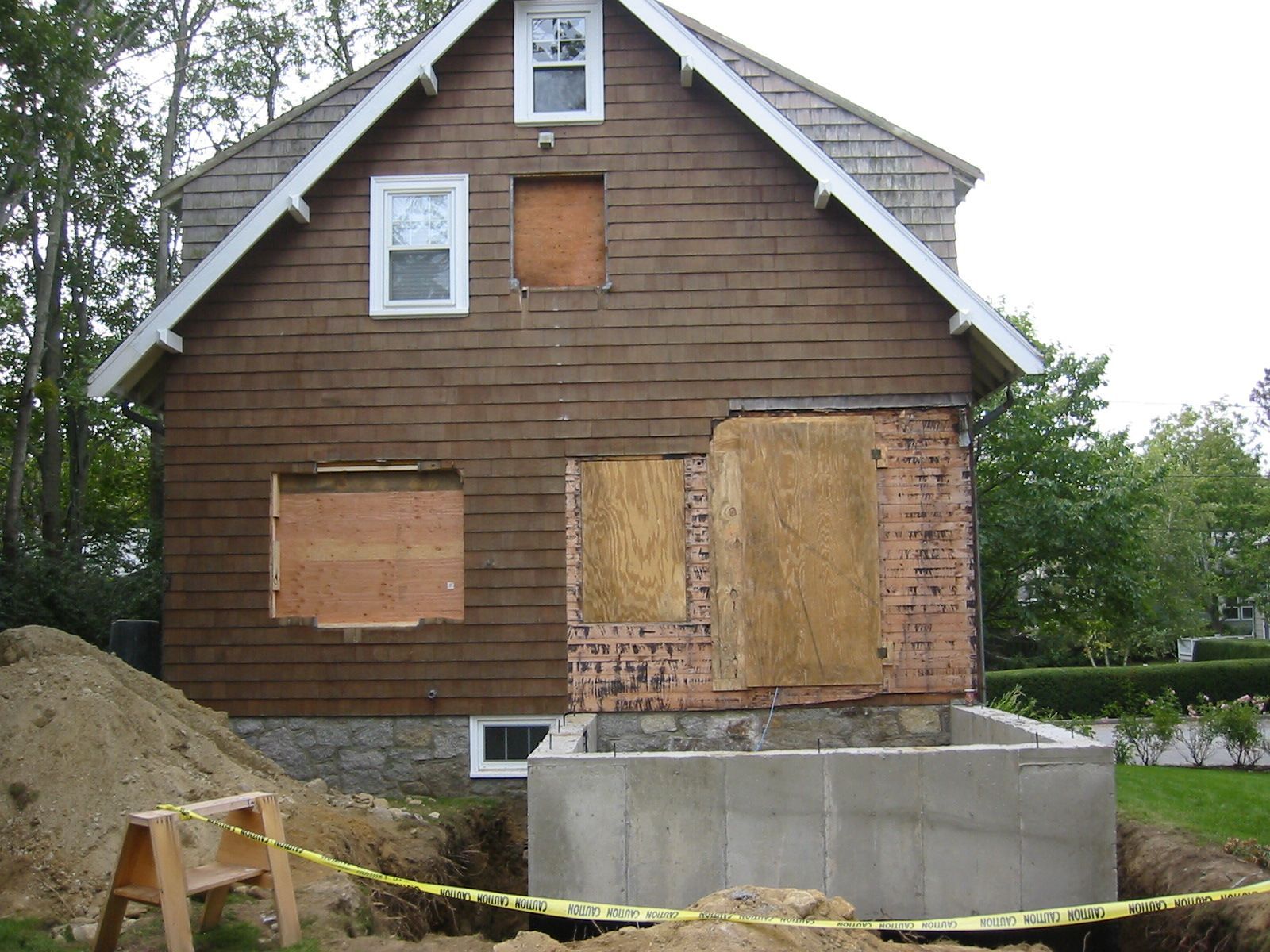 Back of brown house under construction, with missing siding, plywood covering openings, and a new concrete foundation.