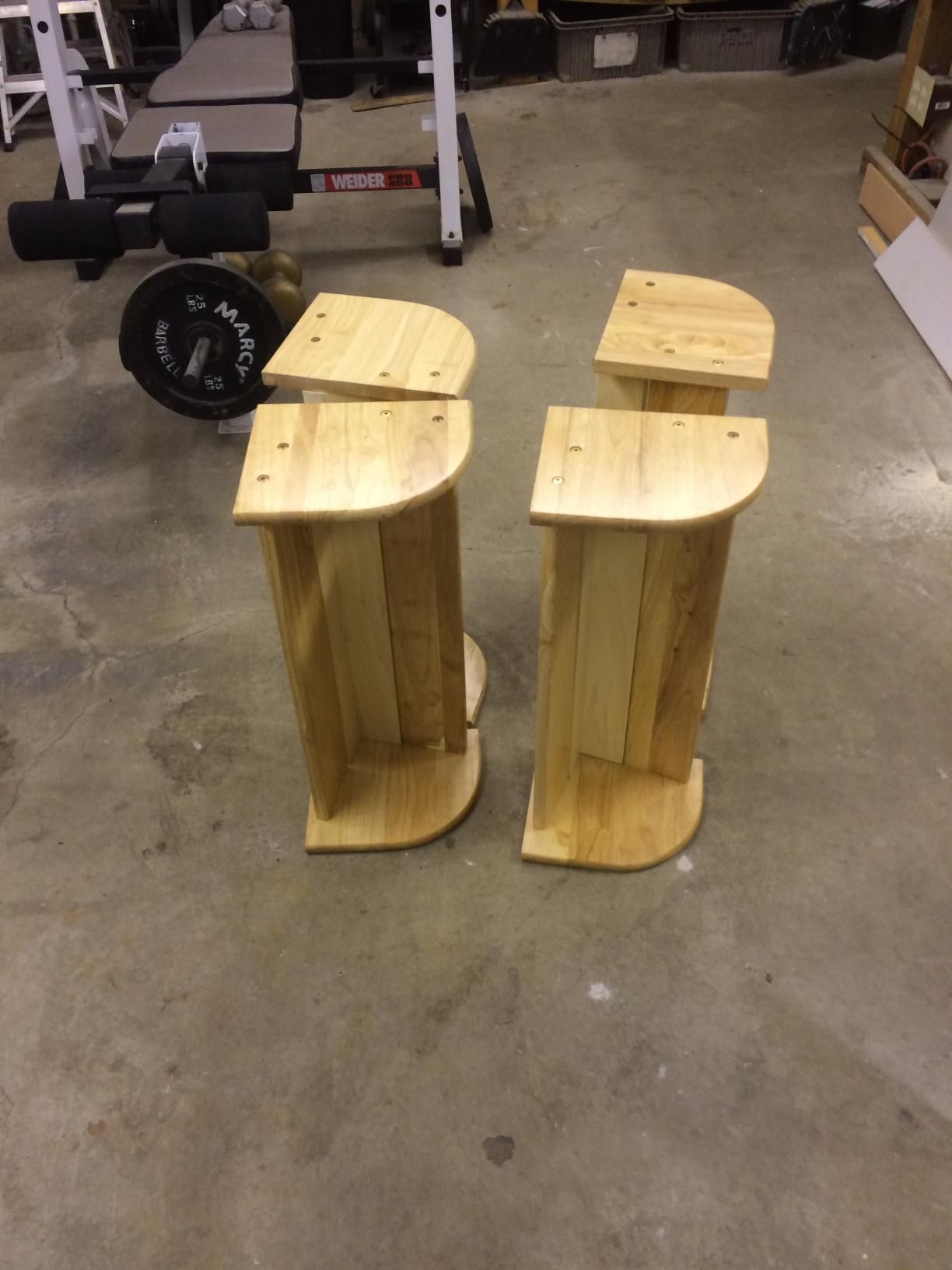 Four wooden, curved-edge speaker stands, unfinished, on a concrete floor, near gym equipment.