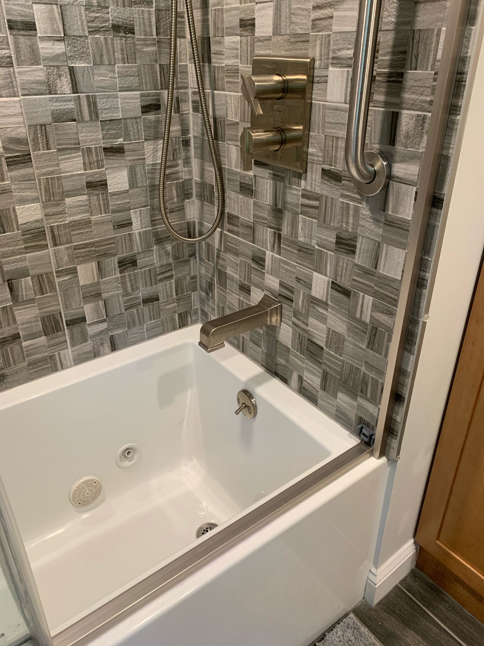 White bathtub with silver fixtures and grey mosaic tile backsplash.