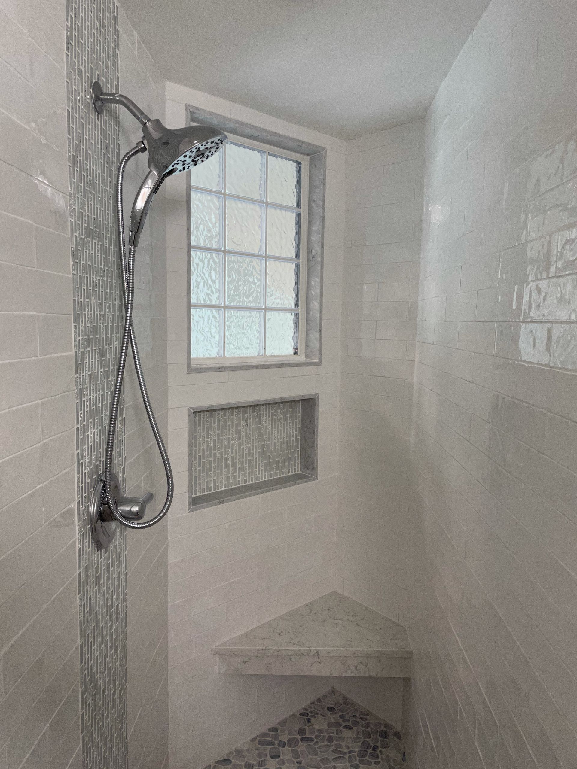 Shower with a window, featuring a chrome showerhead and corner shelf. Tiled in white and grey.