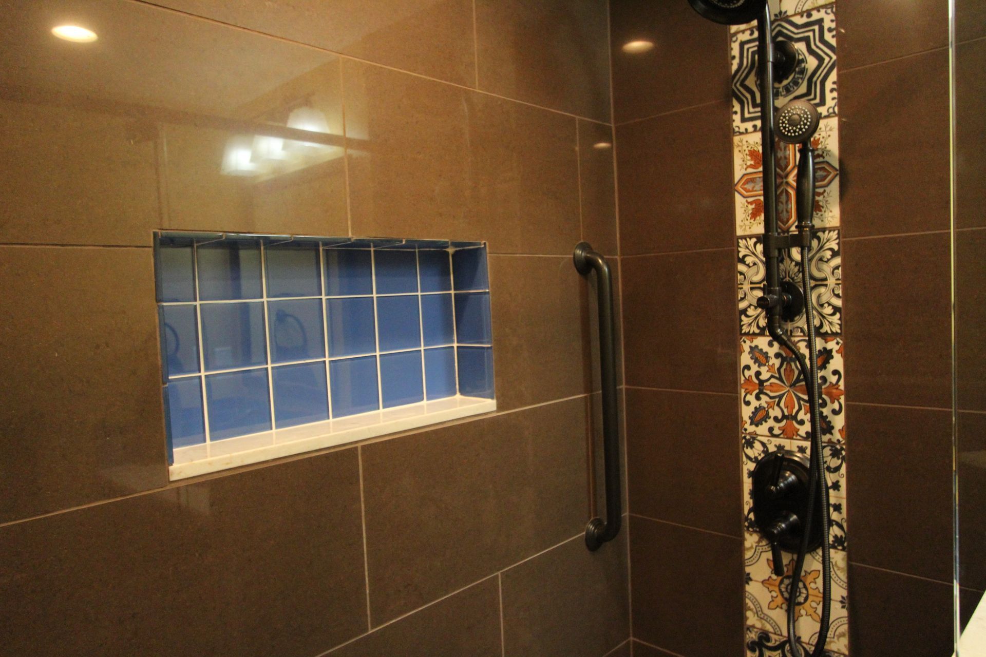 Brown-tiled shower interior with a blue-tiled niche. A decorated shower column and grab bar are visible.