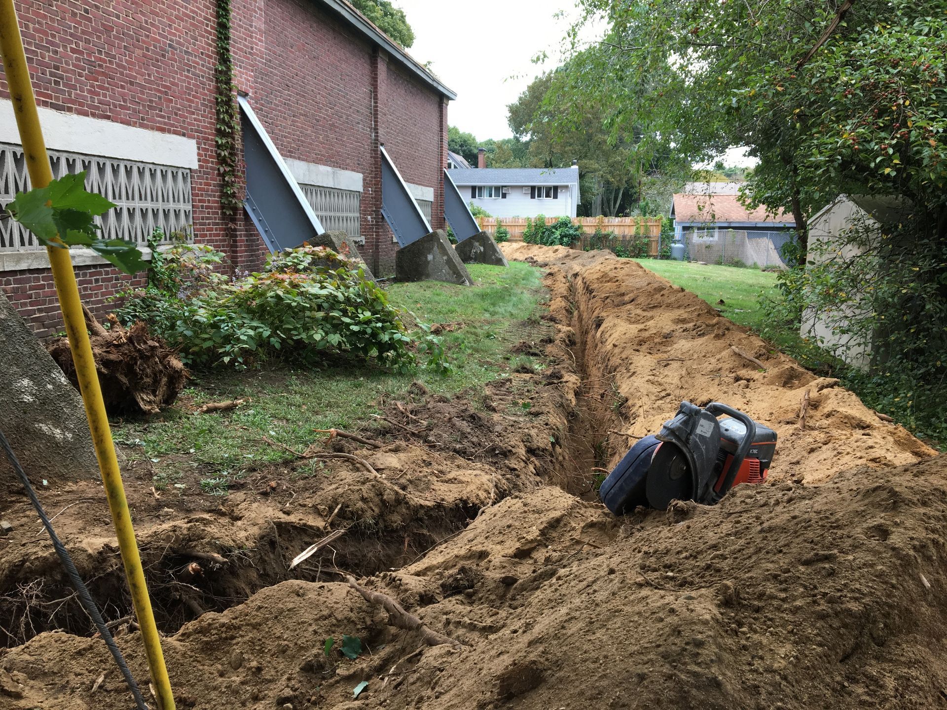 Man digging a trench next to a brick building. Dirt piles and a view of houses in the background.