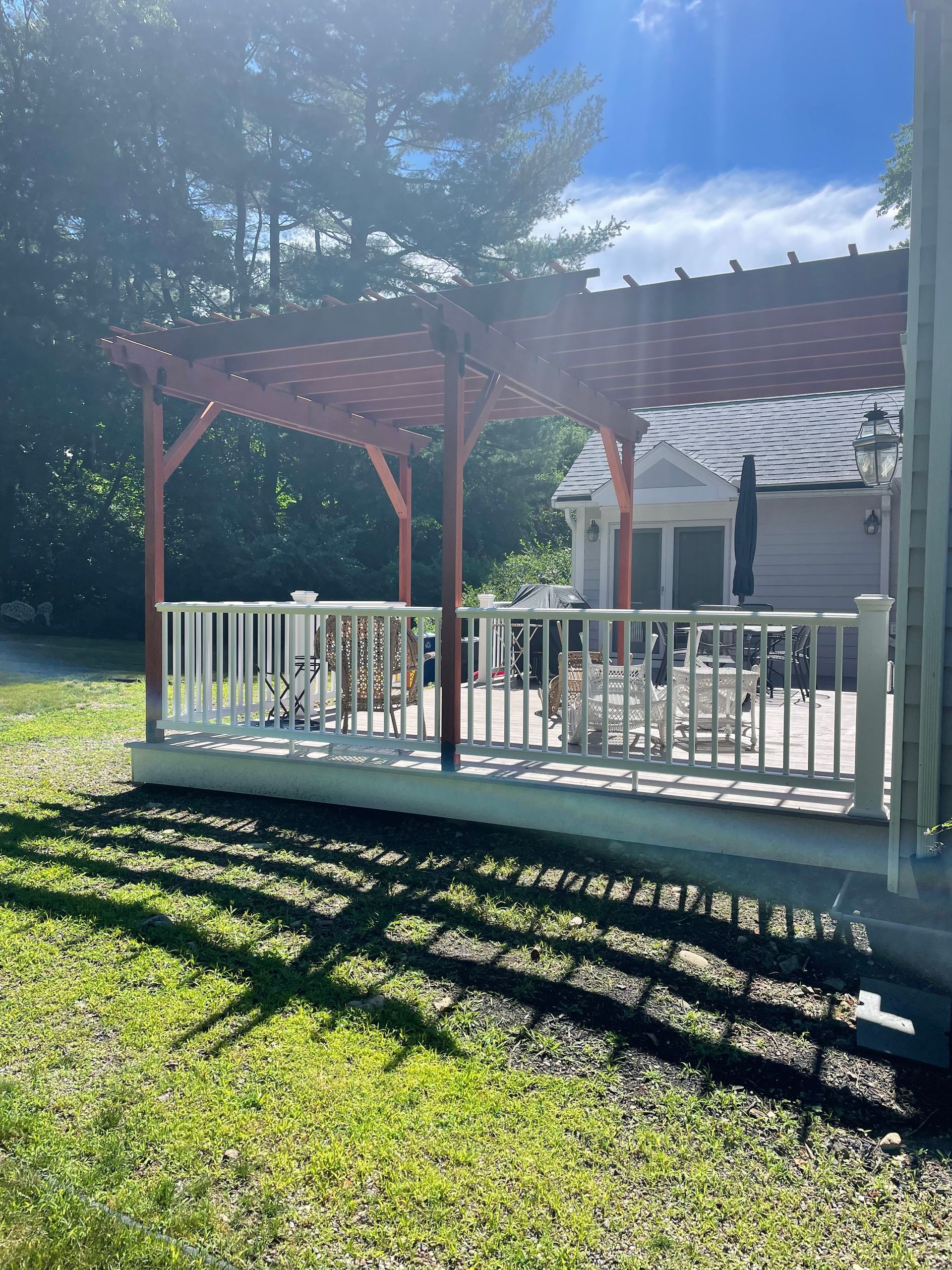 Outdoor patio with a pergola, white railing, and shadows on grass, under a bright blue sky.