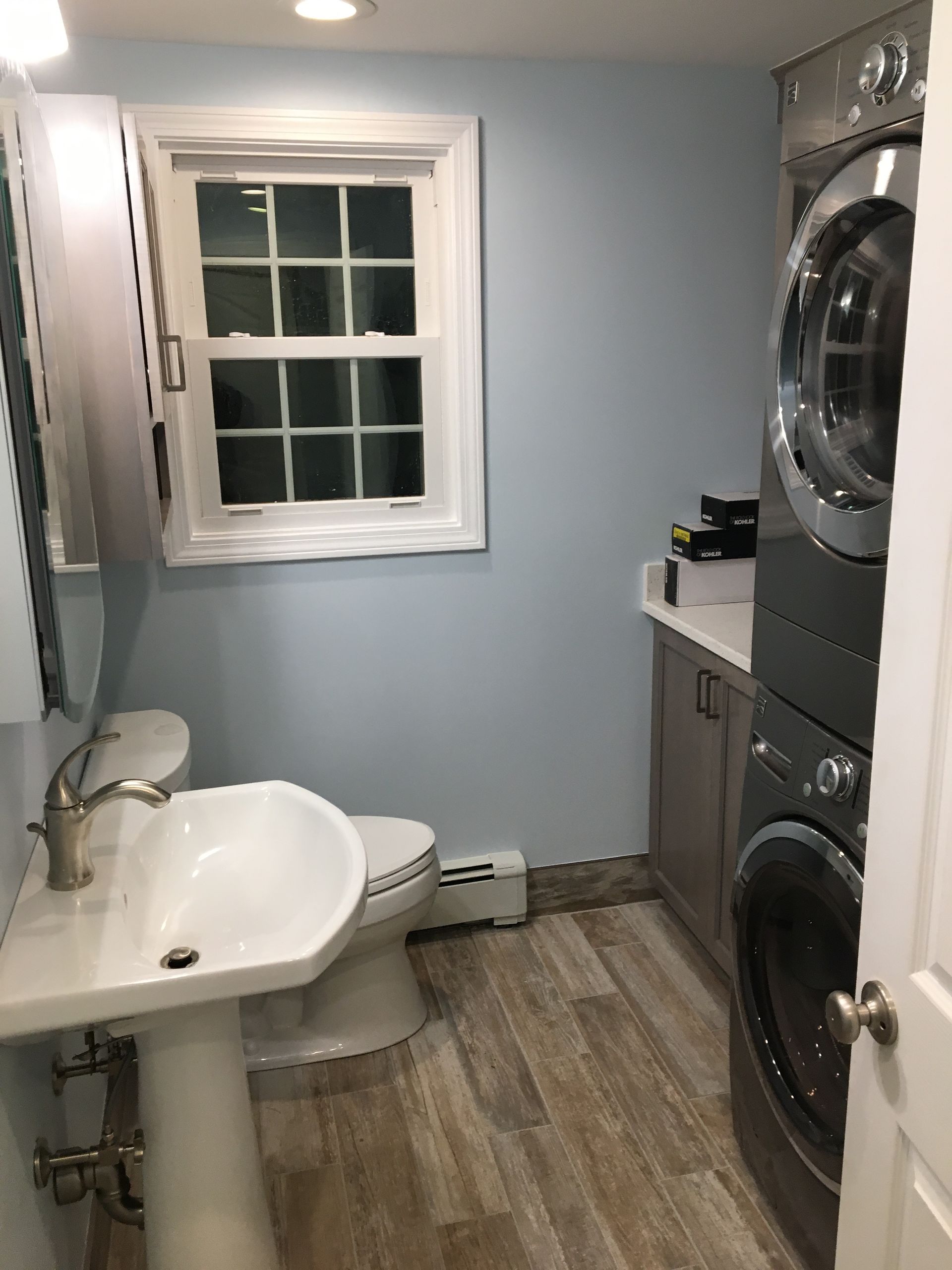 Bathroom with pedestal sink, toilet, window, stacked washer/dryer, light blue walls, and wood-look flooring.