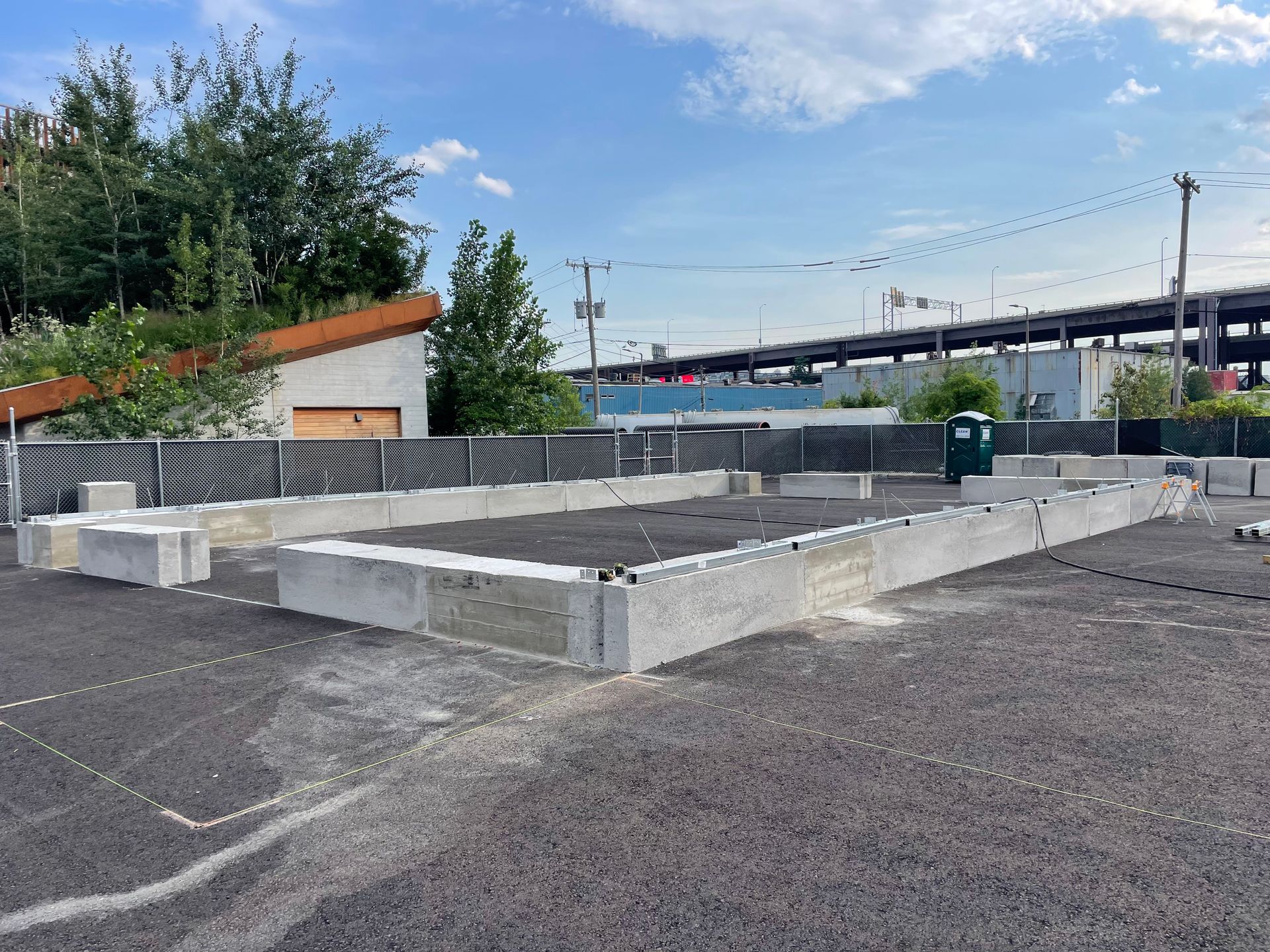 Concrete foundations with a gravel surface, fenced area, and a bridge in the background under a blue sky.