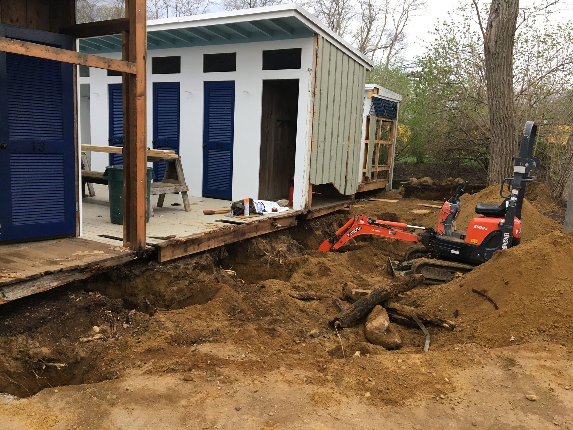 Excavator digging under a small, white and blue building with blue shutters; construction in progress.