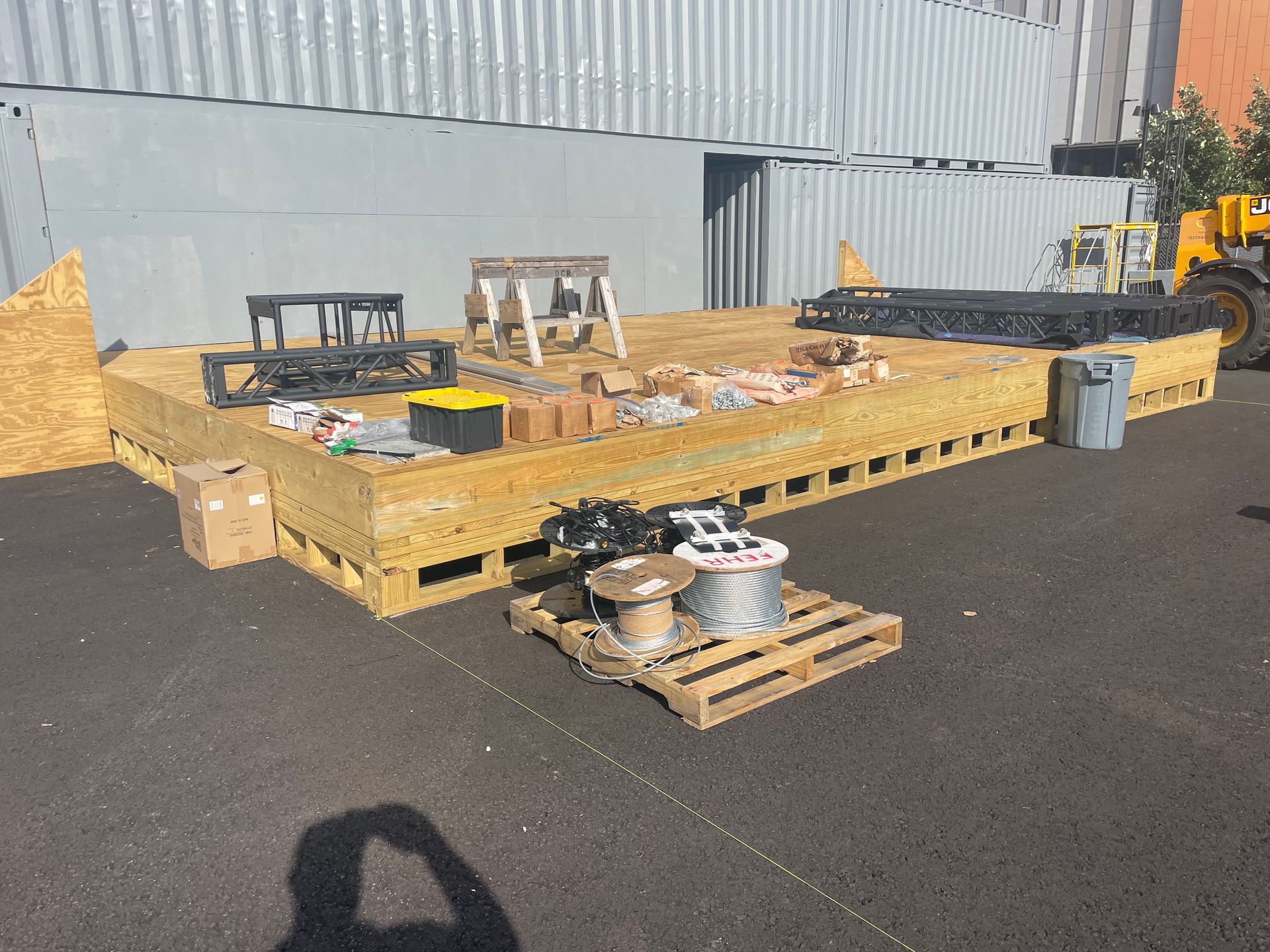 Wooden platform with building materials and equipment, next to shipping containers.