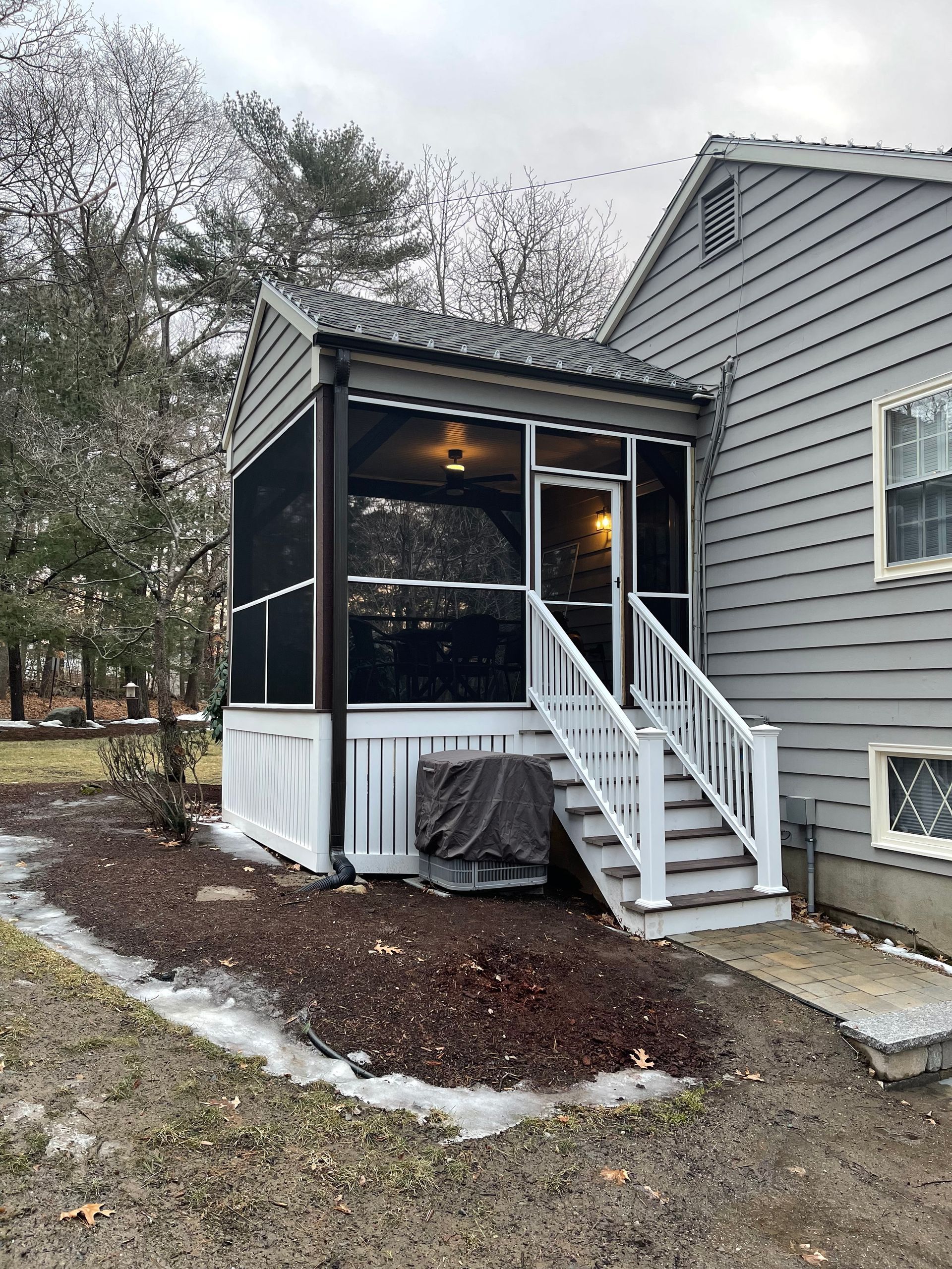 Screened-in porch attached to a gray house, white trim, stairs, and a covered air conditioning unit.