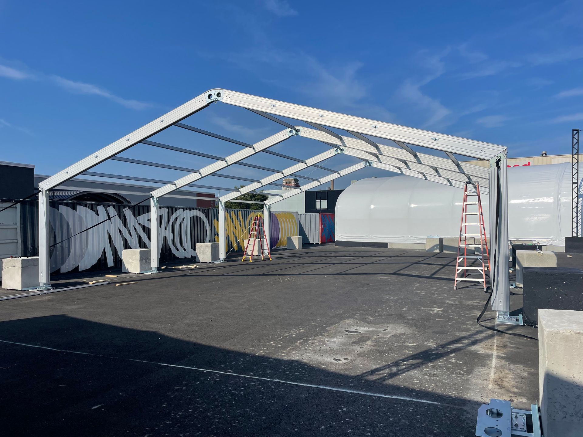 A large, open-sided white tent in a parking lot, with graffiti art on a nearby wall.