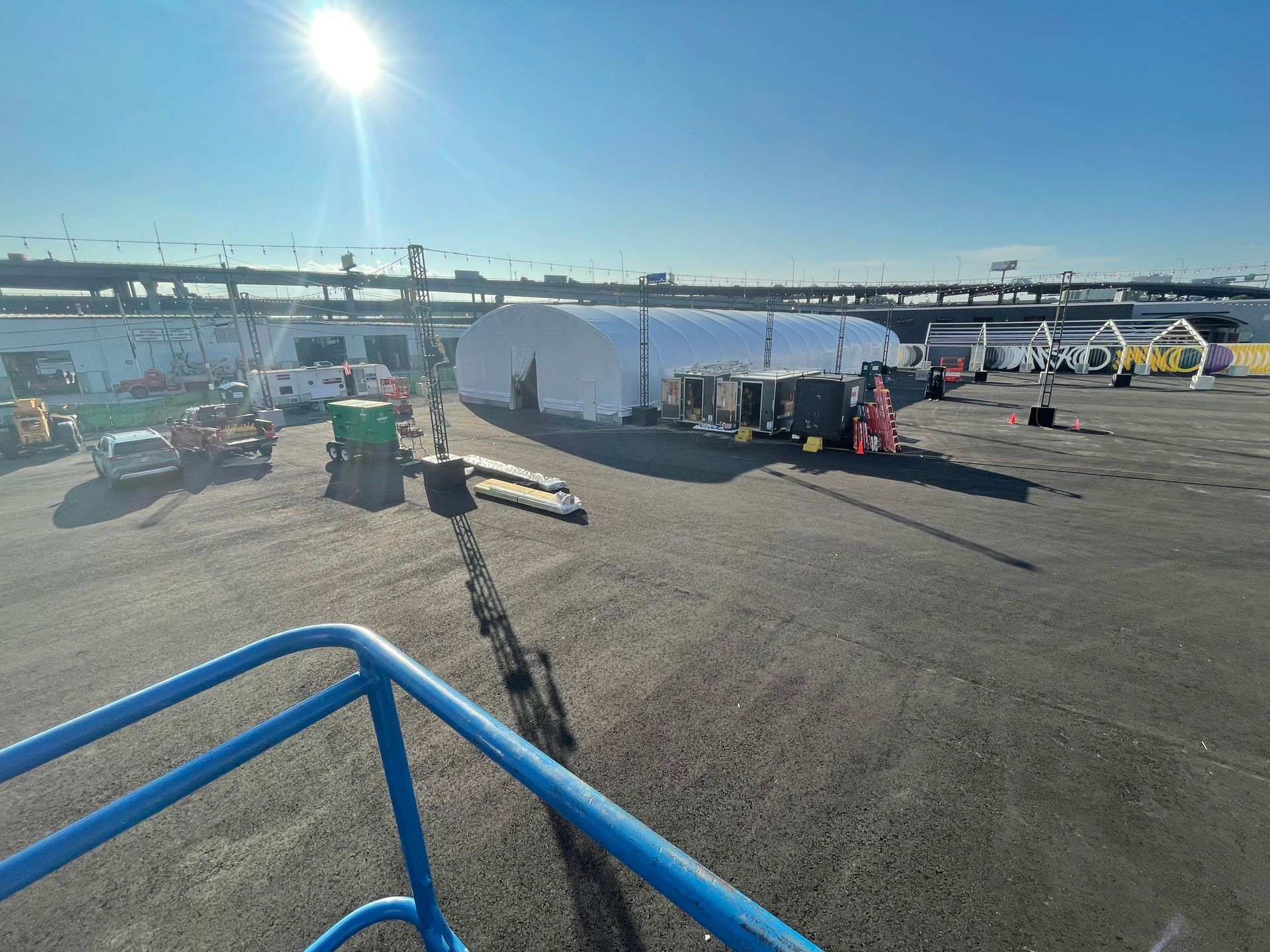 Large white tent in an outdoor lot, equipment and vehicles nearby, bright sunlight.