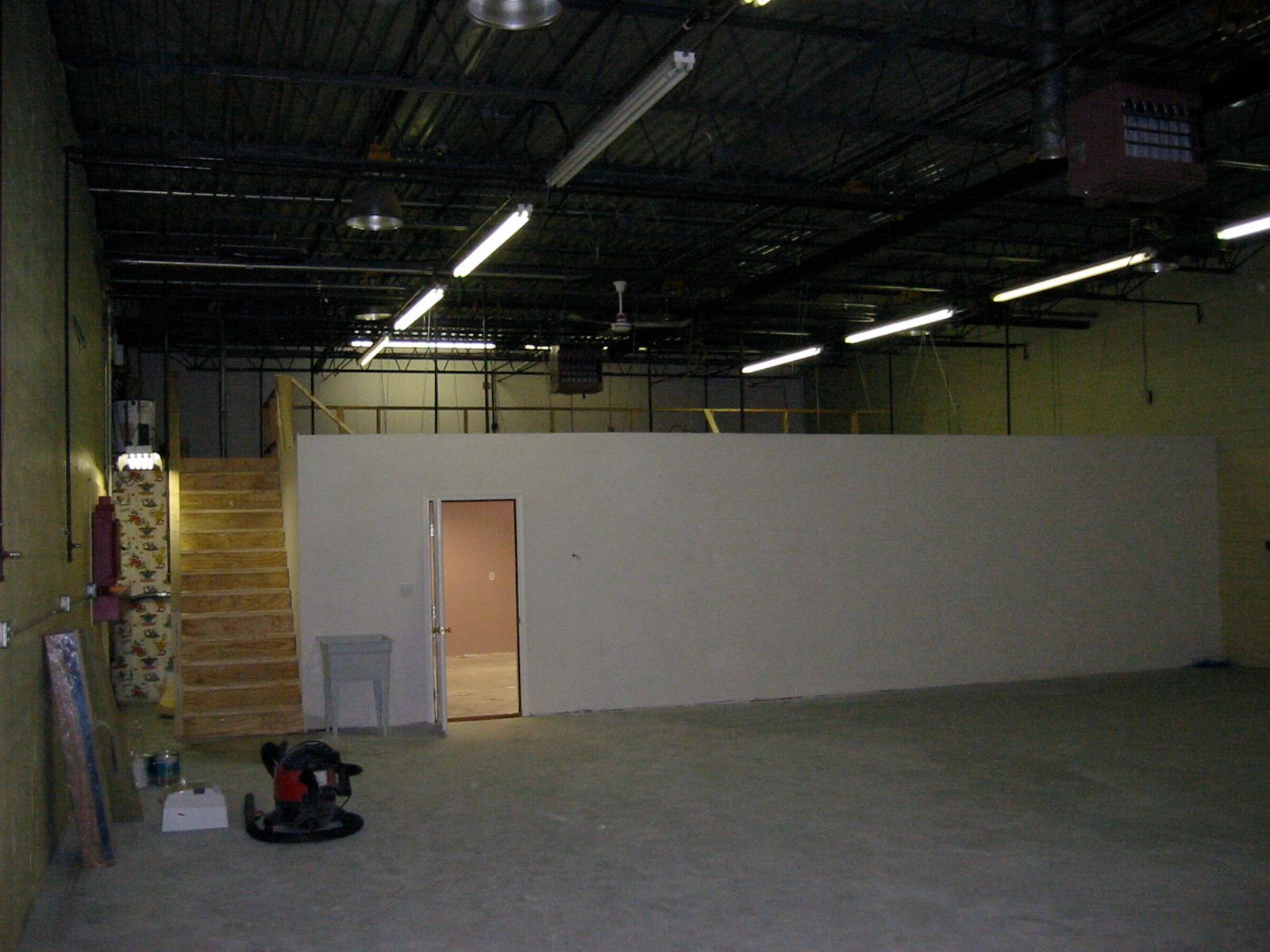 Empty interior of a large warehouse space with a small mezzanine level, staircase, and door.