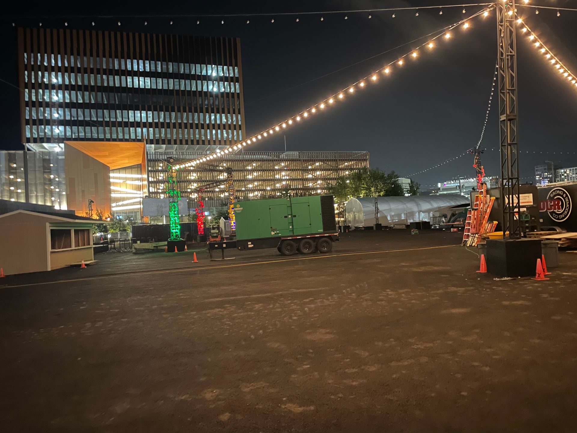 Night scene with string lights, buildings in the background, a green container on a trailer, and a small building to the left.