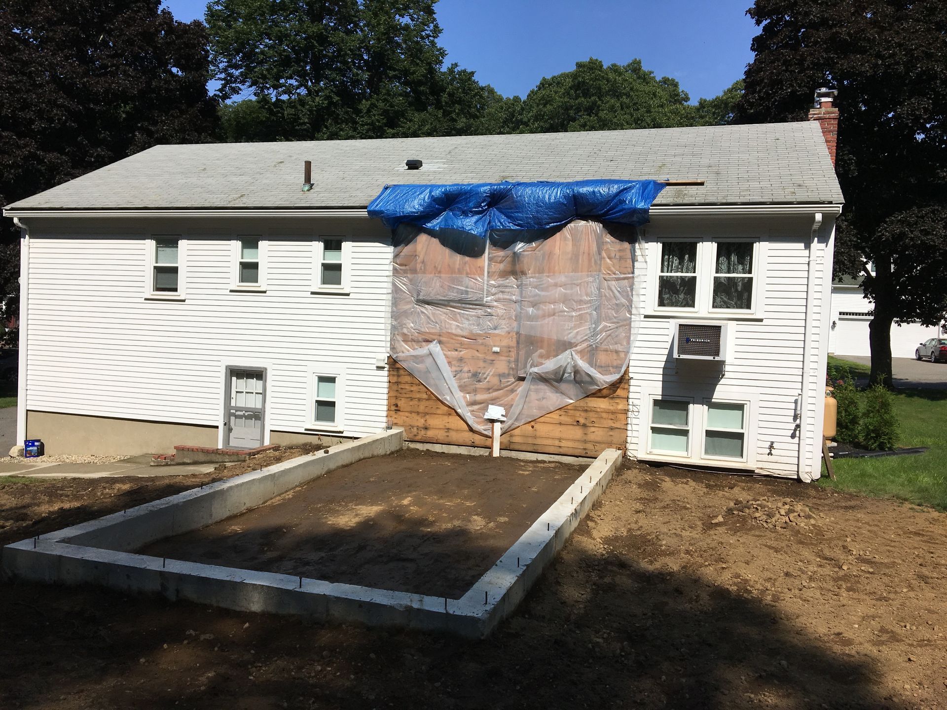 Rear view of a two-story white house with a partially exposed wall covered by a blue tarp. A concrete foundation is in the foreground.