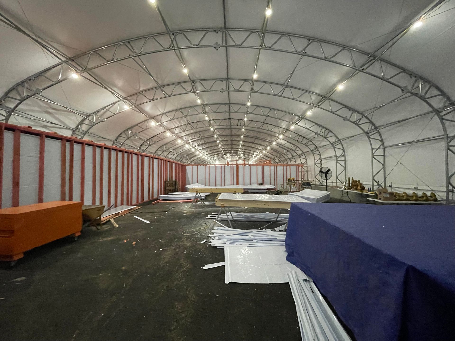 Inside a large, arched tent structure; white roof, metal frame, red and white walls, dark floor, and various supplies.