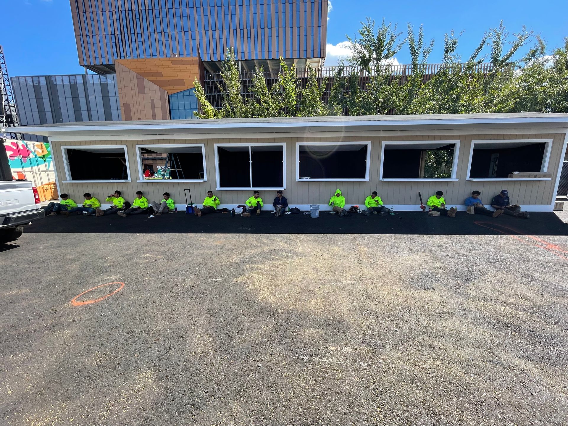 Construction workers in green vests sit in front of a long, low building with windows. Asphalt ground, sunny day.