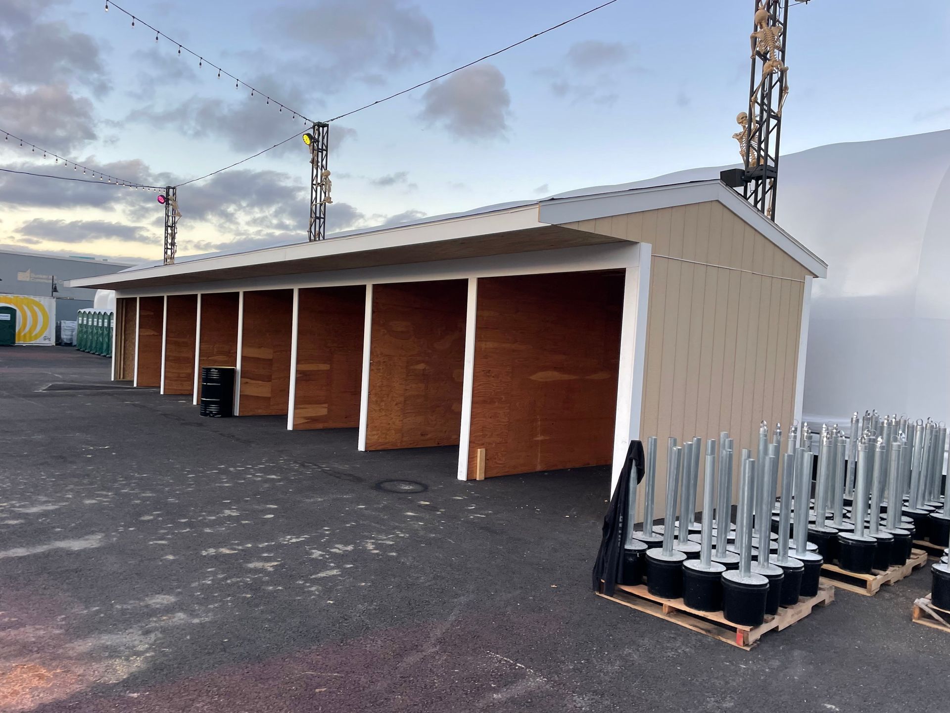 Tan, open-front shed with plywood interior, on asphalt; stacks of black-based silver poles in foreground.