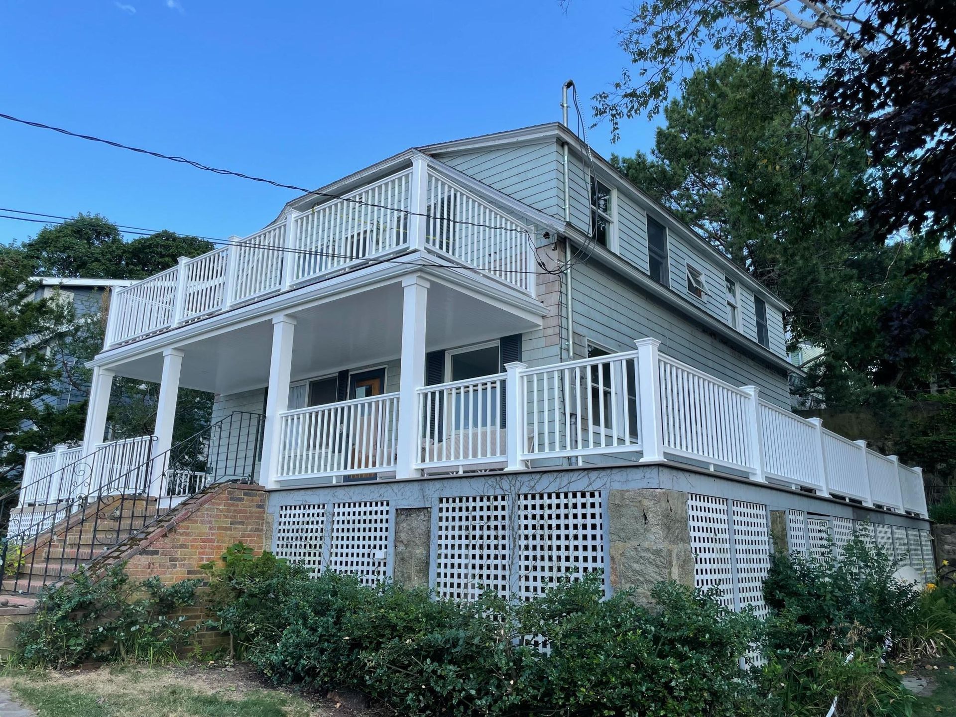 Two-story blue house with white railings and stone foundation, surrounded by greenery and a blue sky.