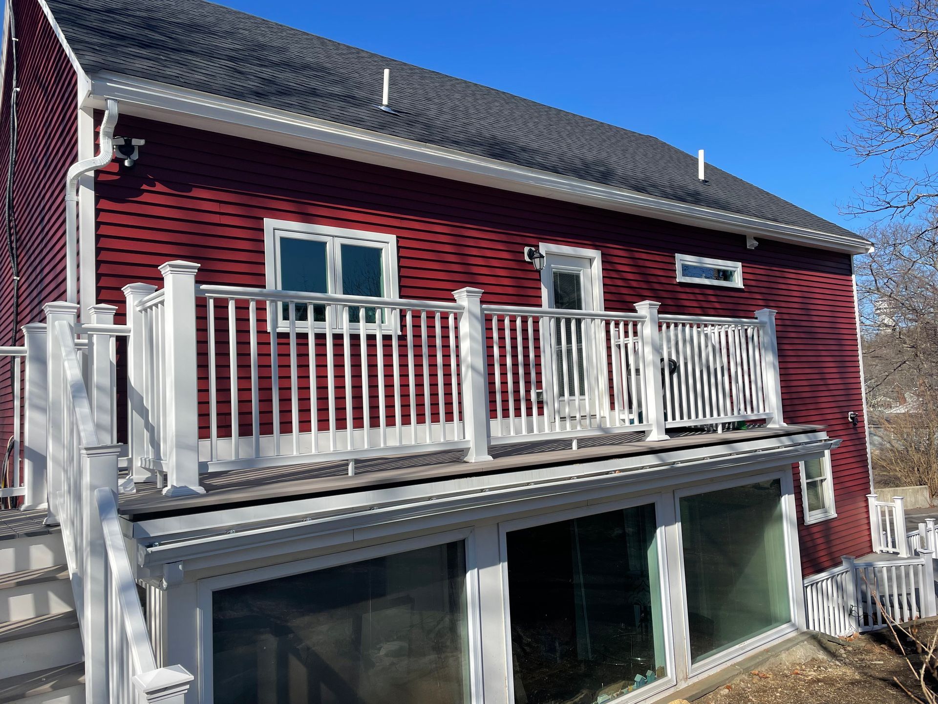 Red house with white deck and stairs, overlooking glass-walled lower level. Clear blue sky.