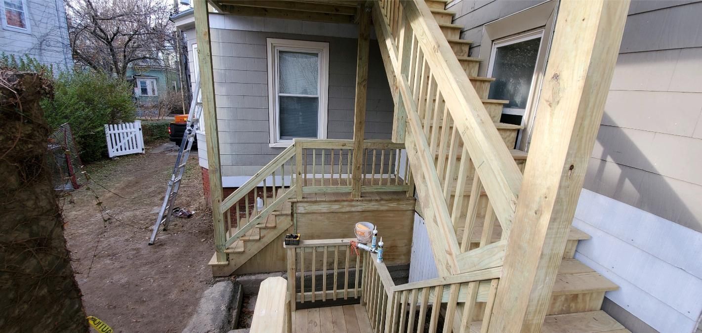 Wooden staircase and porch attached to a gray house. Gravel and a small fence are in the yard.