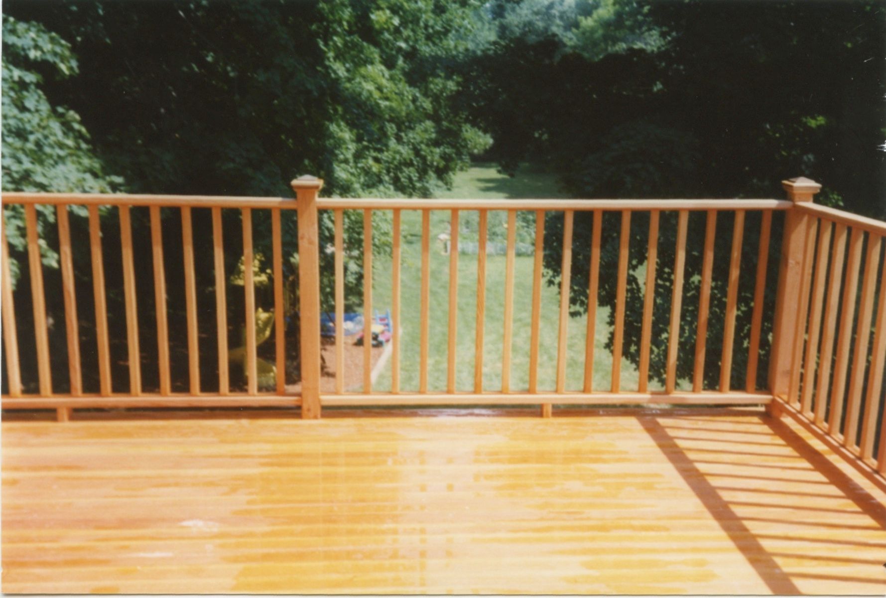 Wooden deck with railing, overlooking a grassy area and trees on a sunny day.