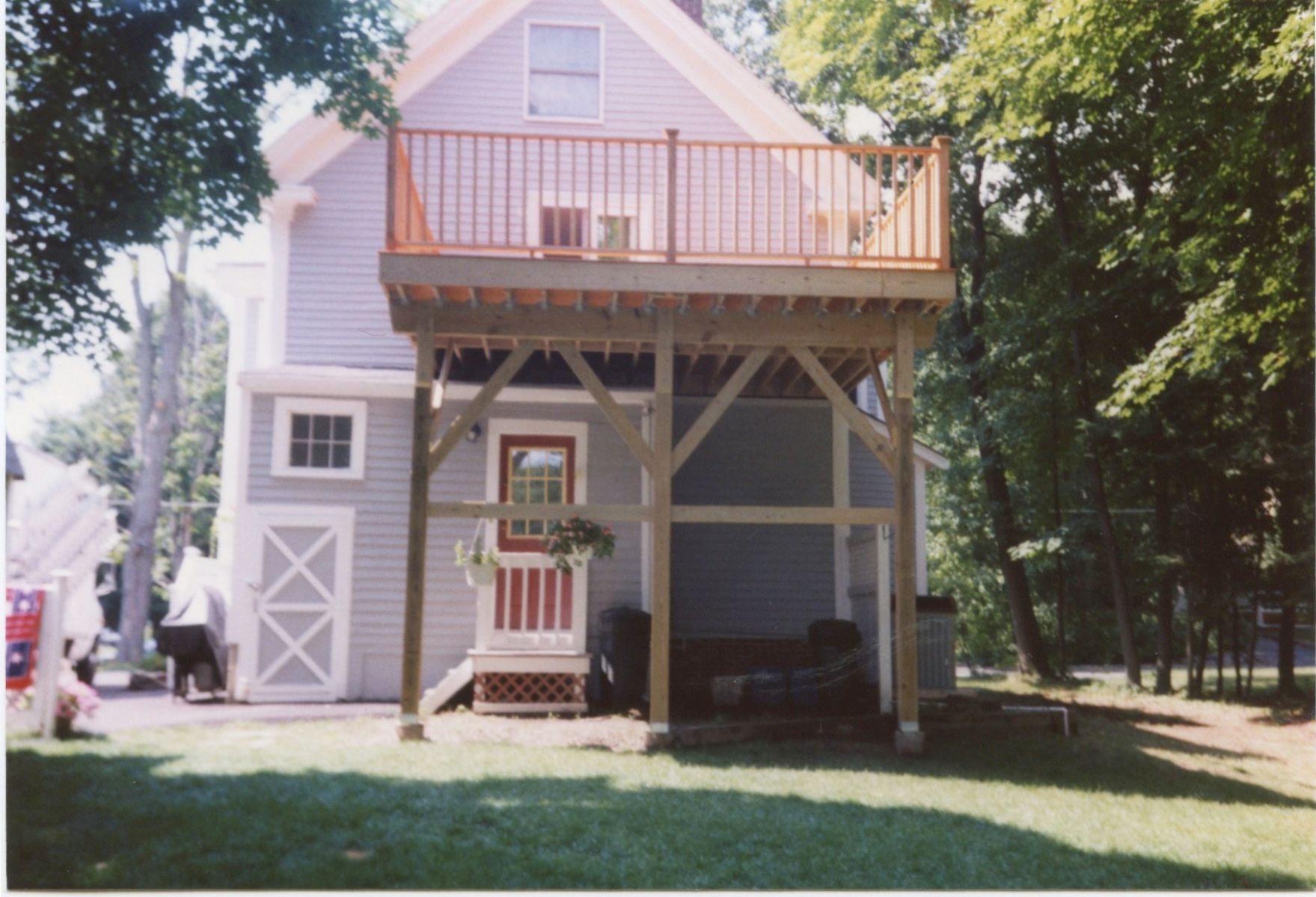 Two-story house with a large wooden deck, supported by posts. Lawn in the foreground; trees on either side.