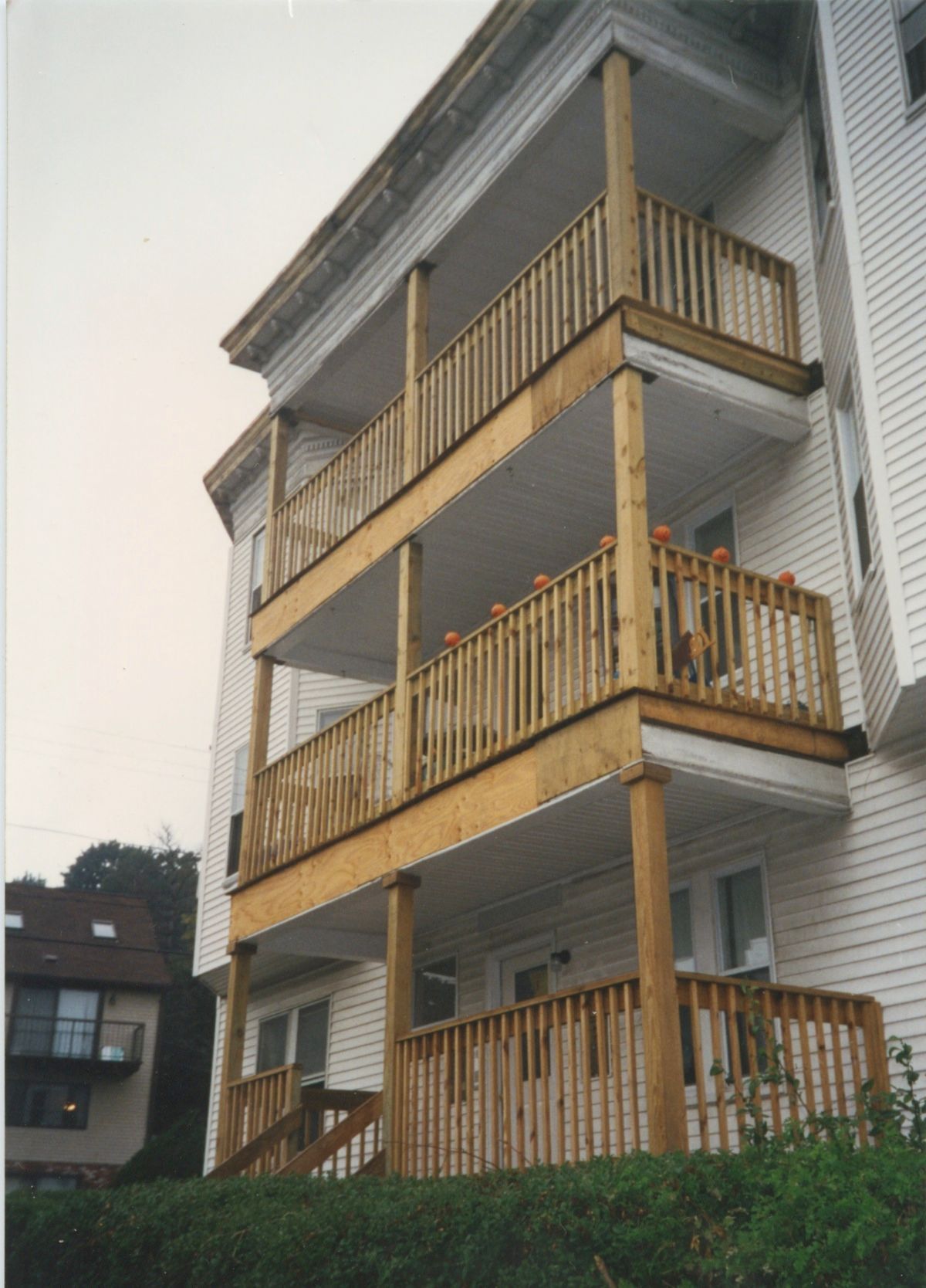 Three-story wooden balconies attached to a white building, with greenery below and a neighboring house visible.