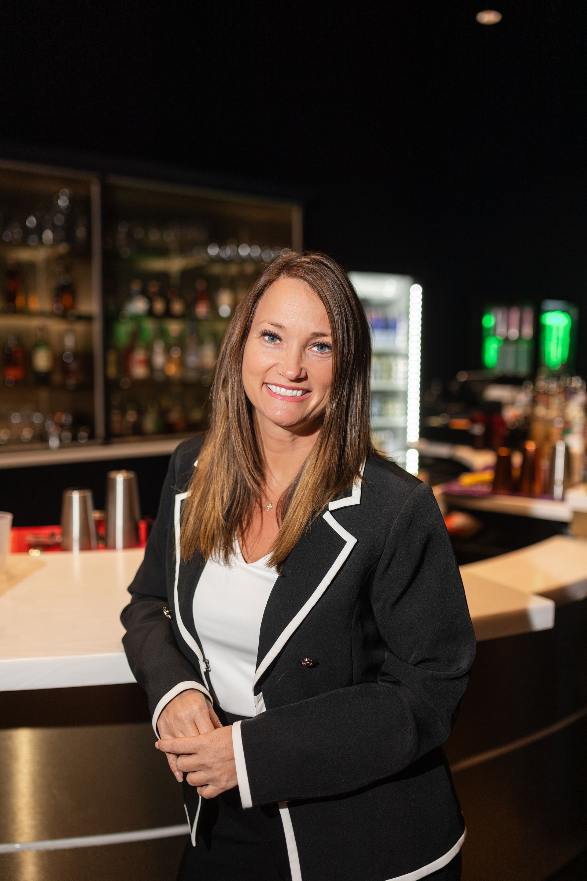Woman leaning on a bar, smiling. Wearing a black blazer with white trim and a white shirt. Bar setting with liquor bottles.