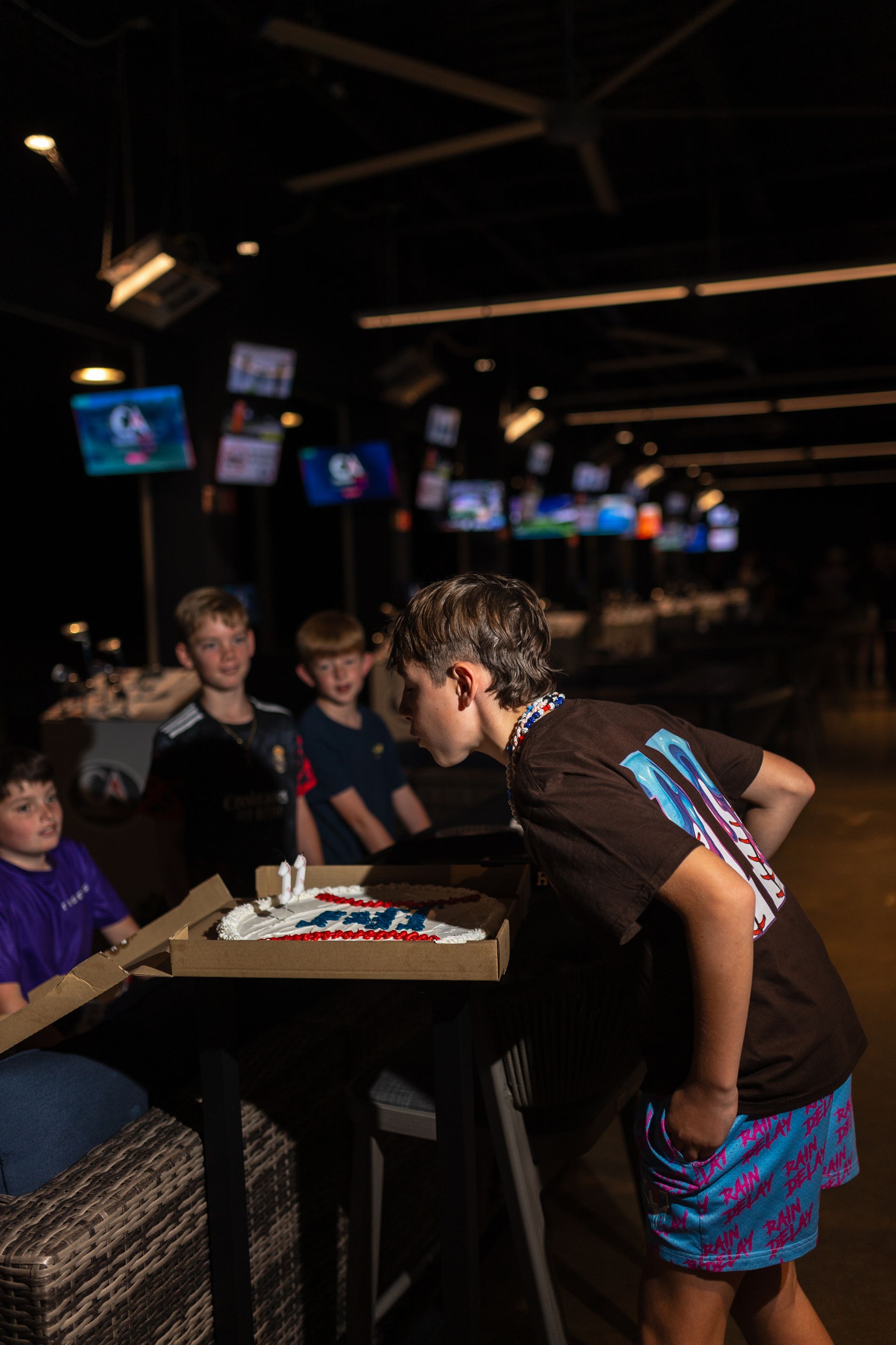 People gathered around a birthday cake at a bowling alley, with a boy leaning over the table to blow out candles.