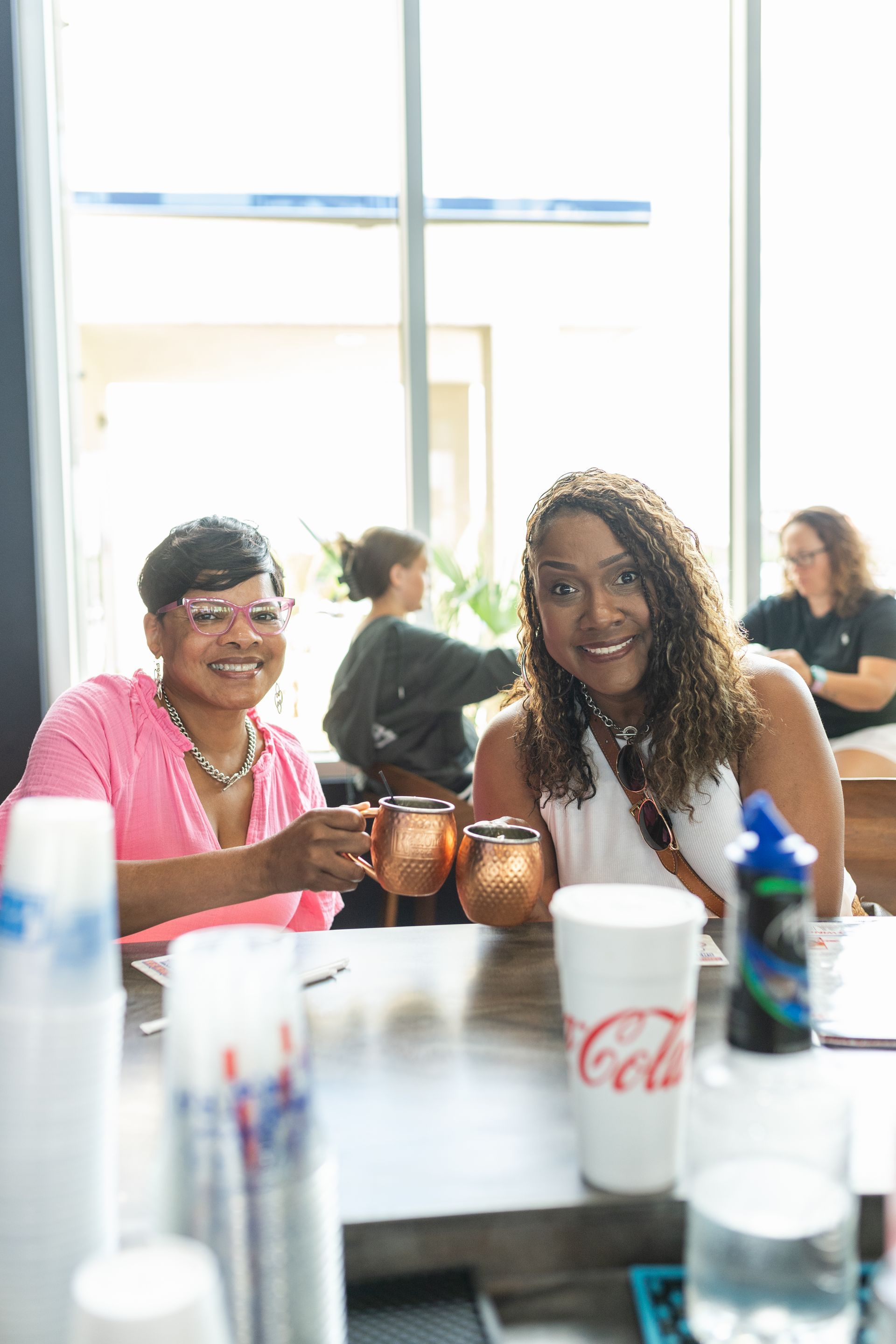 Two women are sitting at a table holding cups of soda.