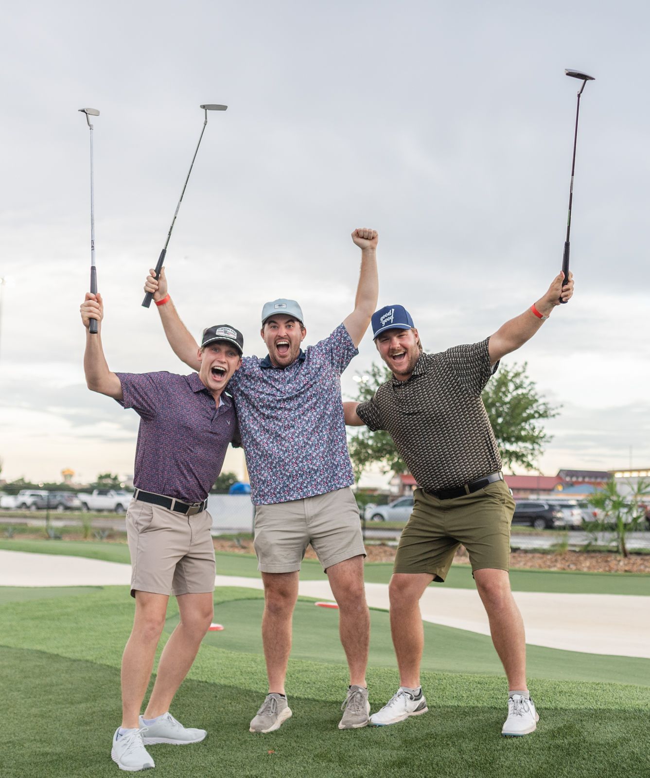 Three men celebrating on a mini golf course, holding putters aloft, arms raised, excited expressions.
