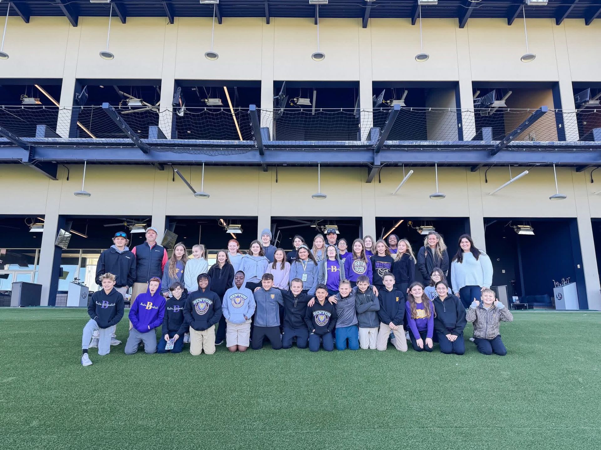 A large group of people smiling and posing for a photo on an outdoor turf field in front of a multi-level golf facility.