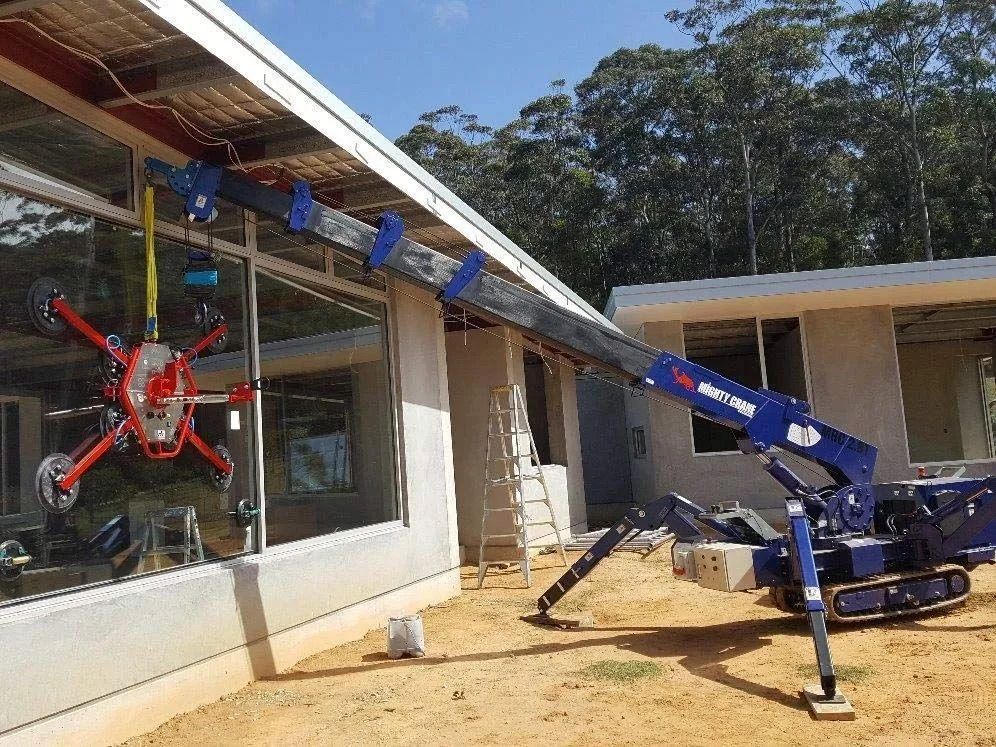 Blue mini-crane lifting red robotic glass handler to install windows on a building under construction — South Coast Glass In South Nowra, NSW
