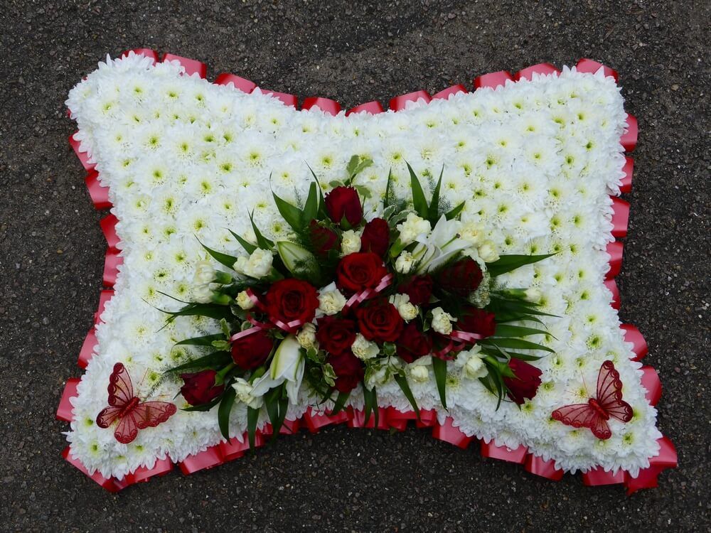 13. 21” white pillow with red ribbon edge, posy of red Roses, cream Dianthus and Lily’s. Butterflies in two corners