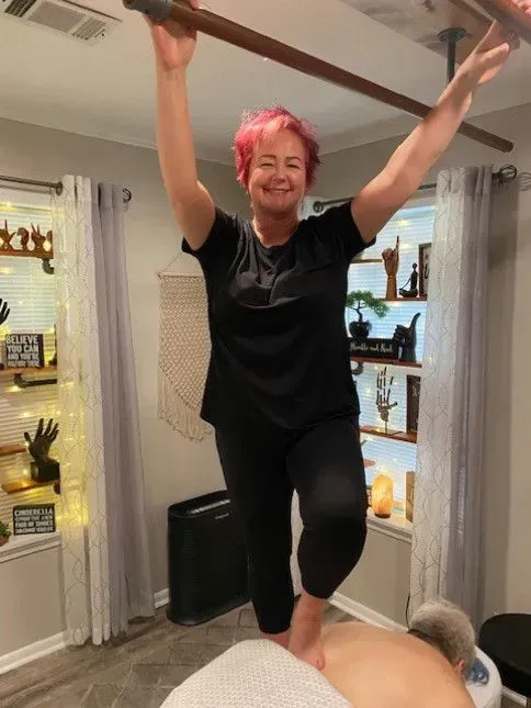 A woman with pink hair is standing on a massage table with her arms in the air.