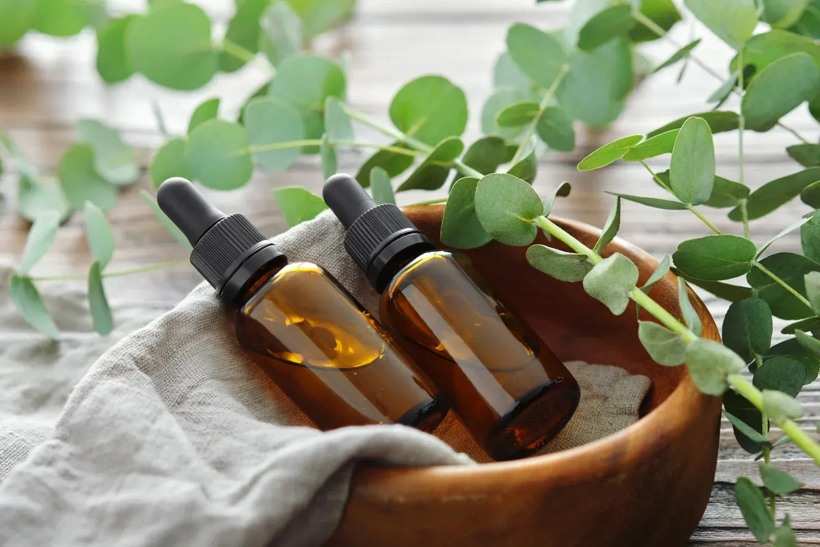 Two bottles of essential oil in a wooden bowl with eucalyptus leaves.