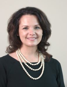 Woman in black top wearing a three-strand pearl necklace, smiling, against a light wall.