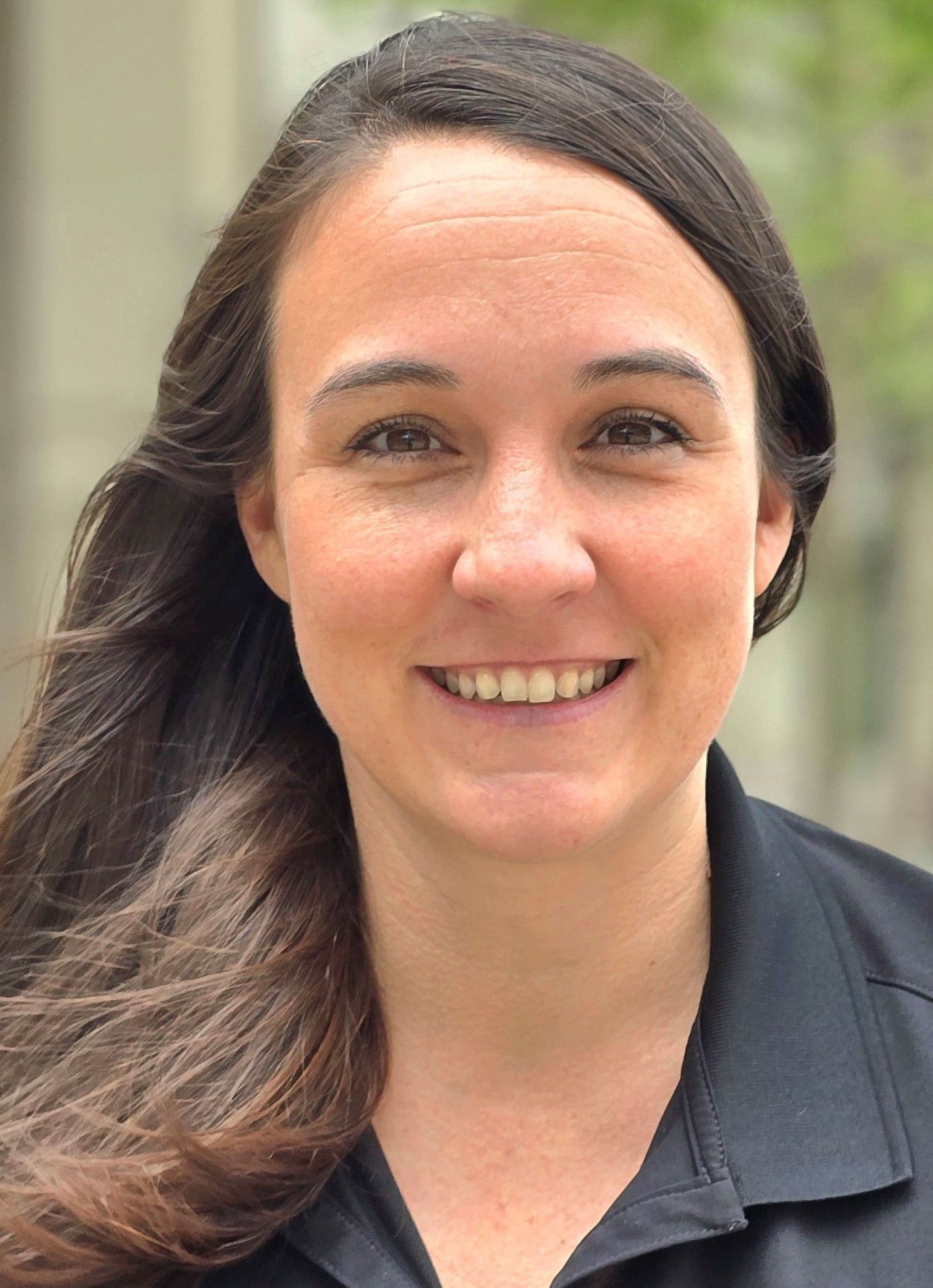 Woman with brown hair smiling, wearing a black shirt, in an outdoor setting.