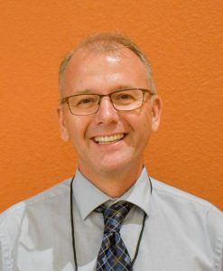 Man in glasses, smiling, wearing a blue shirt and patterned tie. Against an orange background.