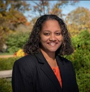 Woman with curly hair smiles, wearing a black blazer and orange top, outdoors with trees in the background.