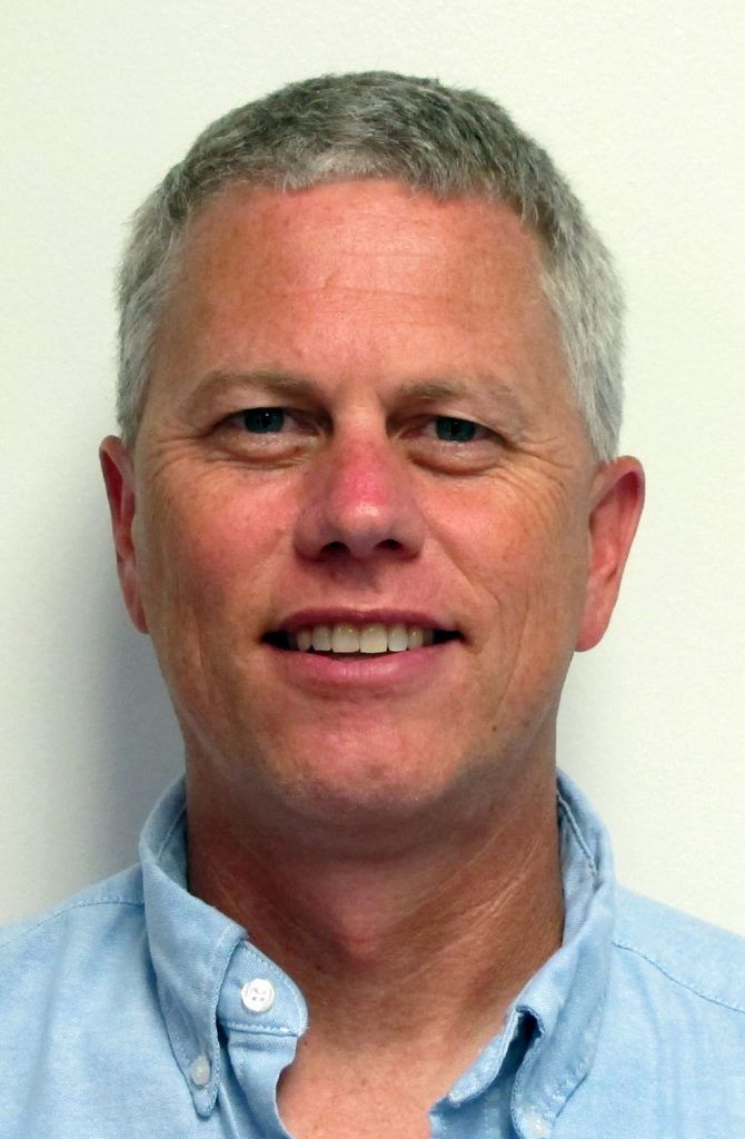 Man with gray hair and blue shirt smiling at the camera, against a white wall.
