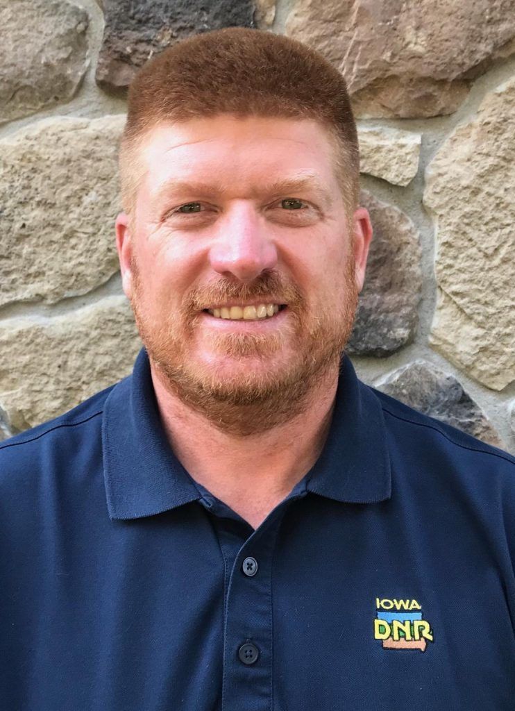 Man with reddish-brown hair, smiling in a blue Iowa DNR polo shirt, in front of a stone wall.