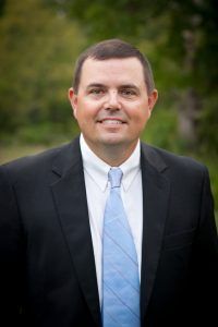Man in a black suit and blue tie smiles, set against a blurred outdoor background.