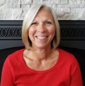Woman with short blonde hair smiles, wearing a red shirt, in front of a fireplace.