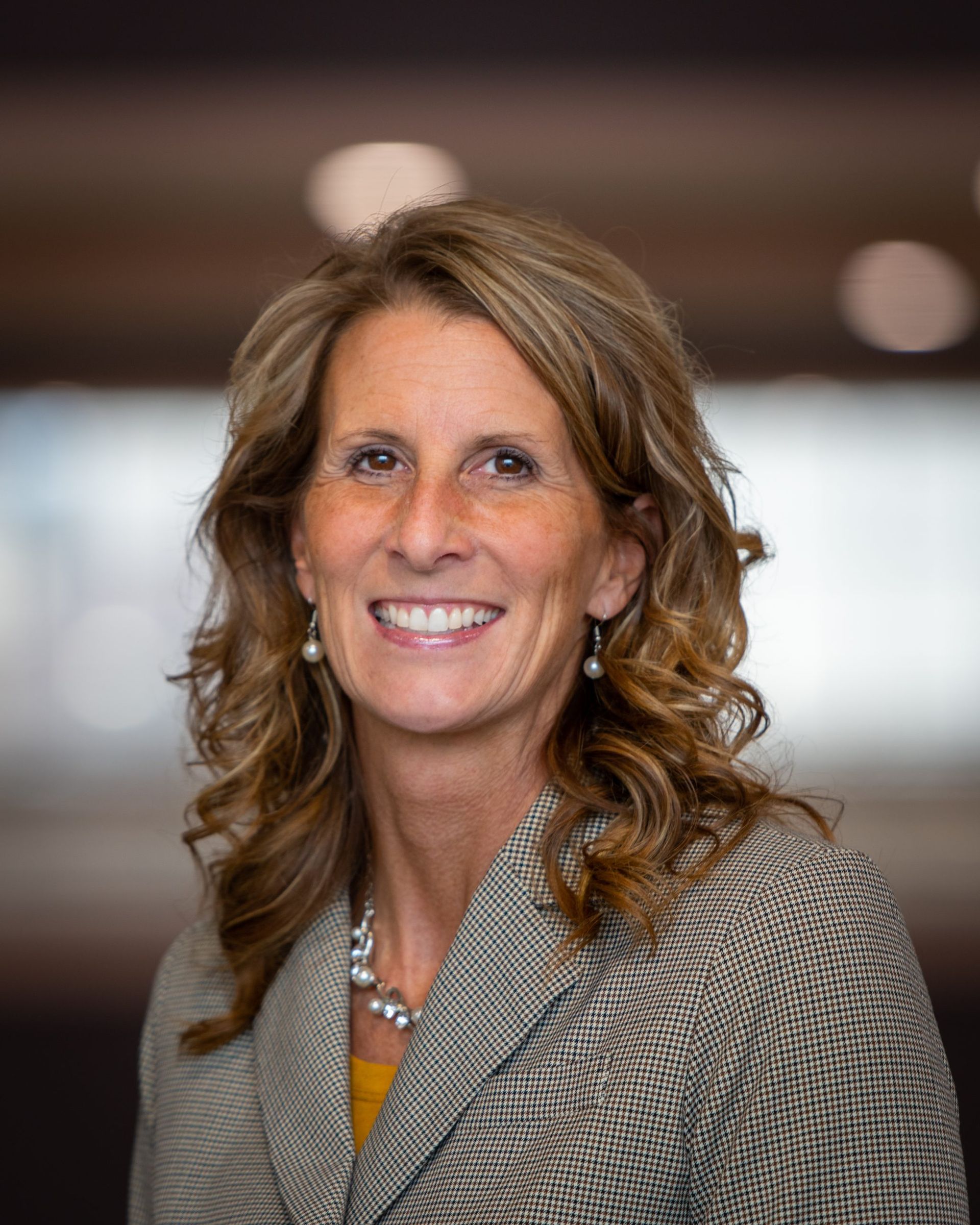 Woman with light brown hair smiles, wearing a patterned blazer and pearl necklace.