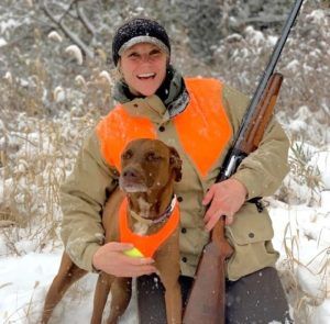 Woman and dog in hunter orange in snow, holding a shotgun.