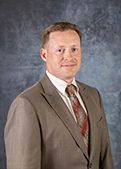 Man in a tan suit, tie, smiling, posed against a mottled blue background.